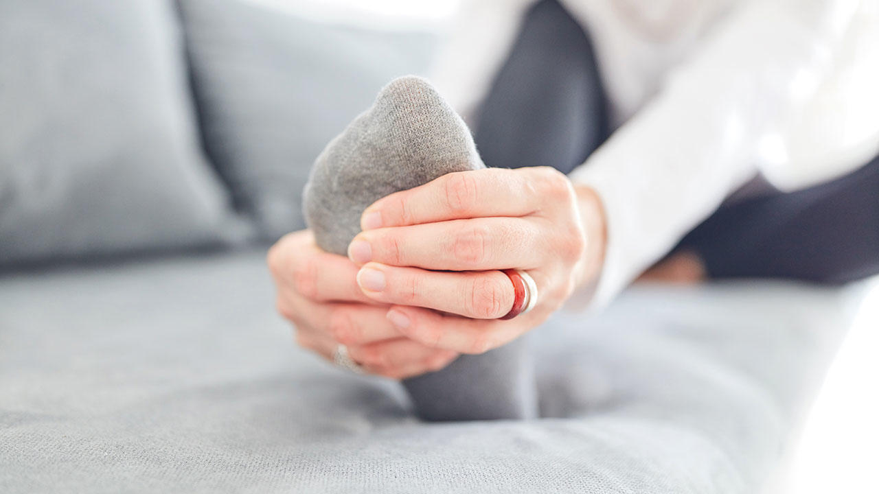 Femme assise sur son canapé, elle se penche en se tenant le pied à deux mains.