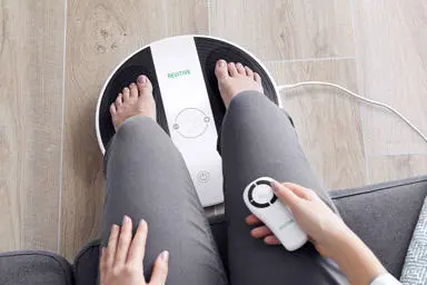 Image of womans legs, with her feet on the prohealth device with the remote control in her hand 